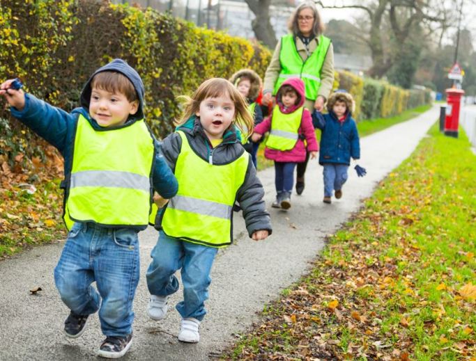 Early years children wearing high visibility vests walking down a pavement accompanied by an adult